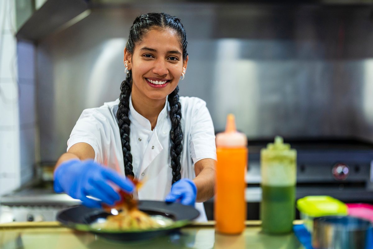 A smiling kitchen trainee in blue gloves plates up a dish