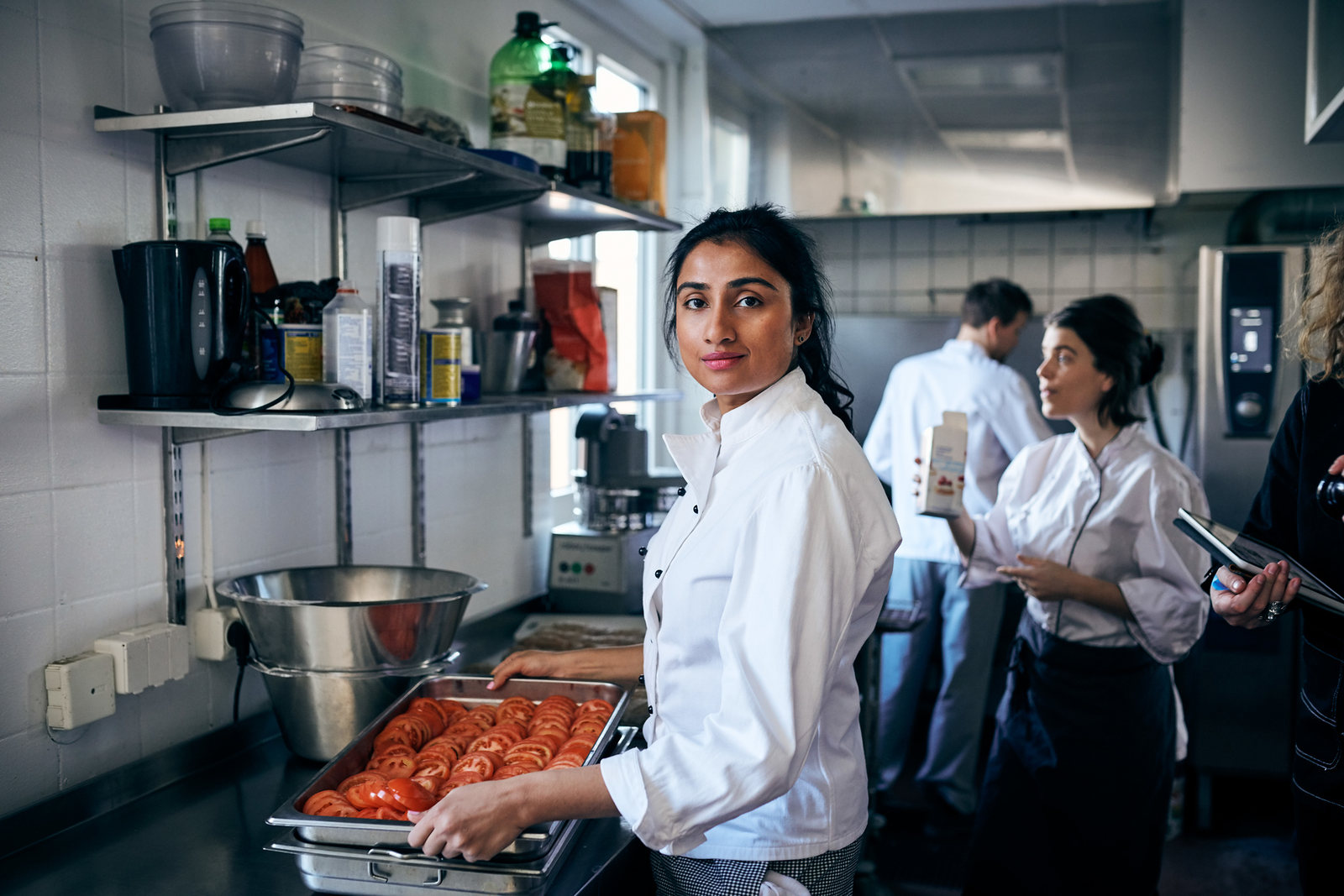 A chef holds a tray of sliced tomatoes in a busy restaurant kitchen while colleagues work behind her